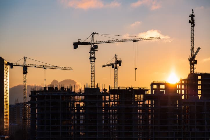 Lots of tower cranes build high-rise residential building in evening time. Steel frame structure in sunlight. Industrial background with sun flare. Yellow sunset sky.